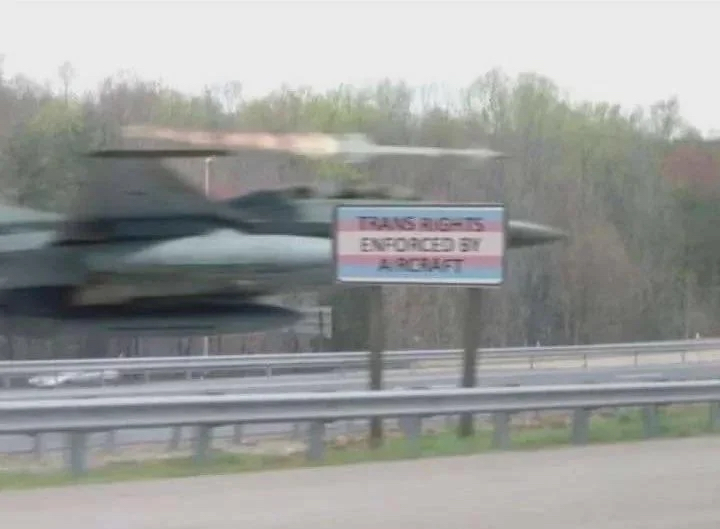 a fighter jet flying extremely low above a highway. the highway has a sign that says trans rights enforced by aircraft, with the trans flag as background. the jet is launching a missile. the image is blurry, especially the jet and missile.